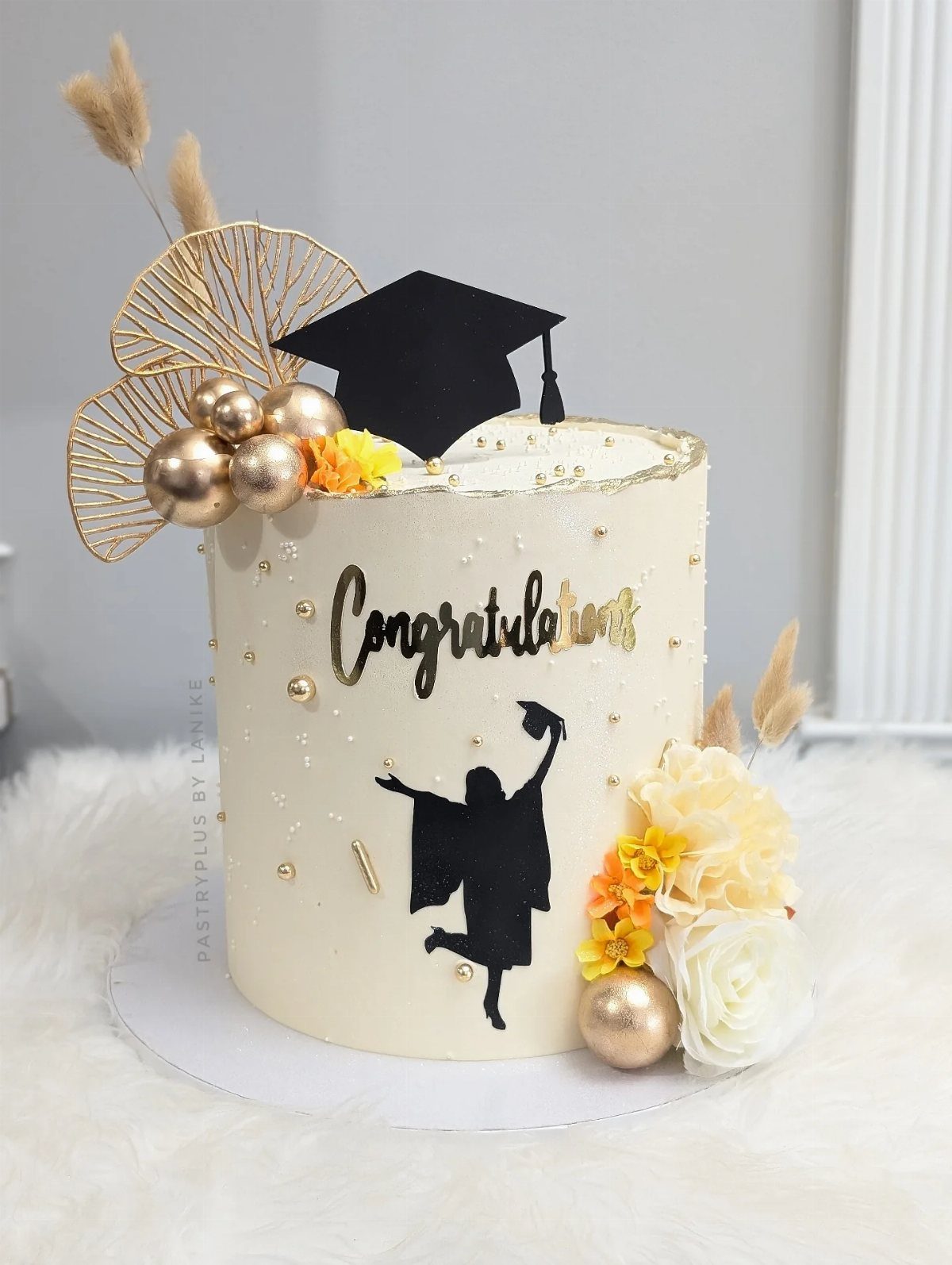 White graduation cake with gold congratulations text, black cap topper, and floral accents.