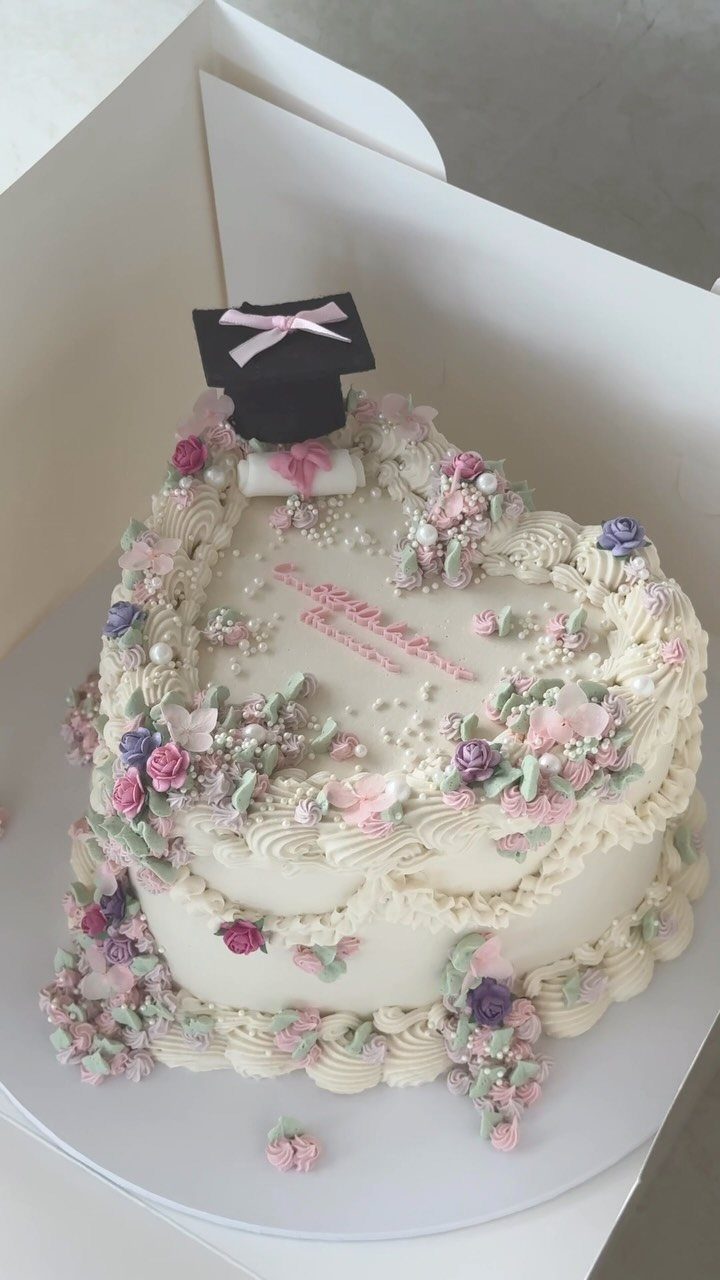 White graduation cake decorated with pink and purple frosting flowers and a cap.
