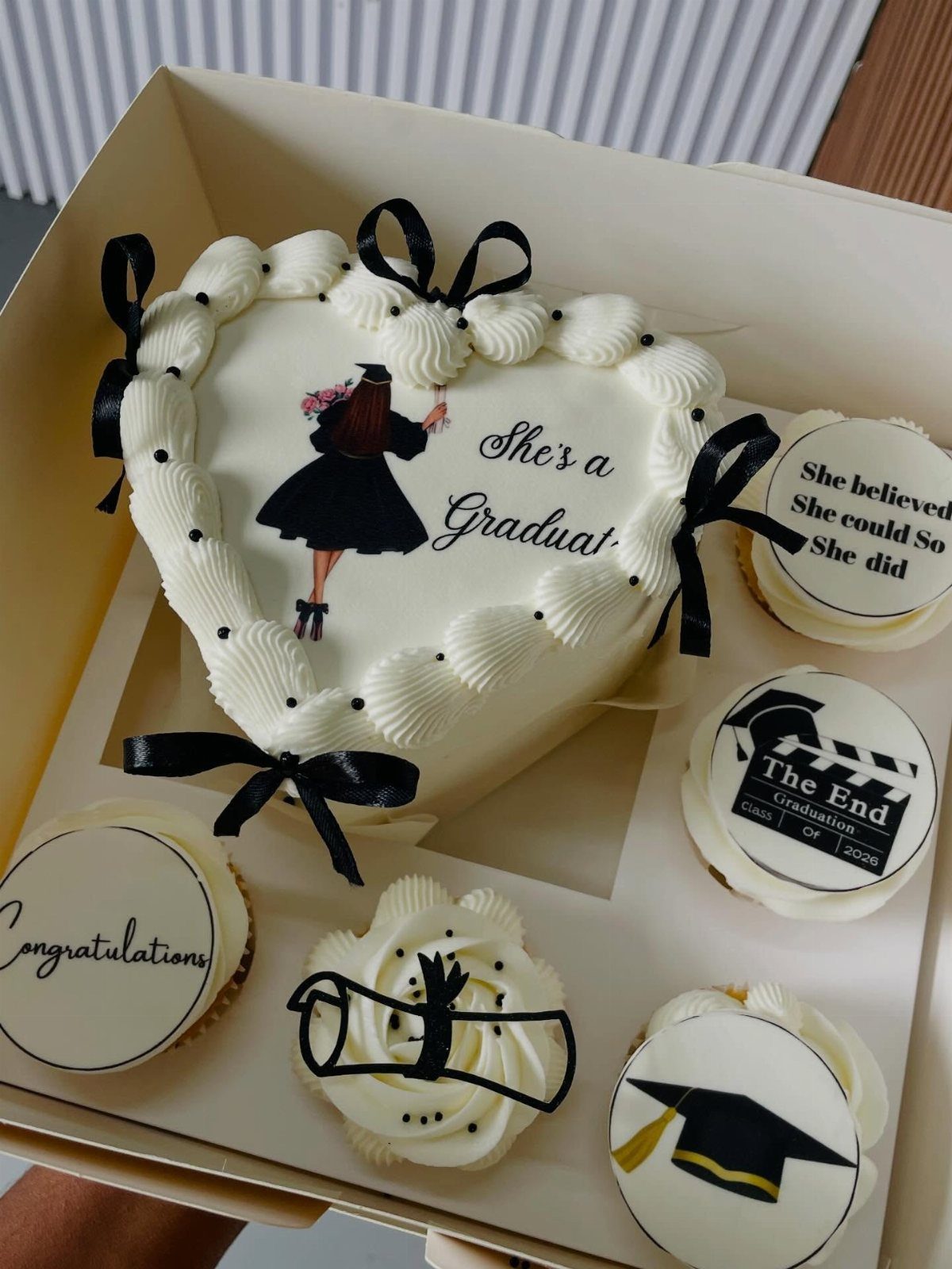 Heart-shaped white graduation cake with black bows and matching themed cupcakes in box