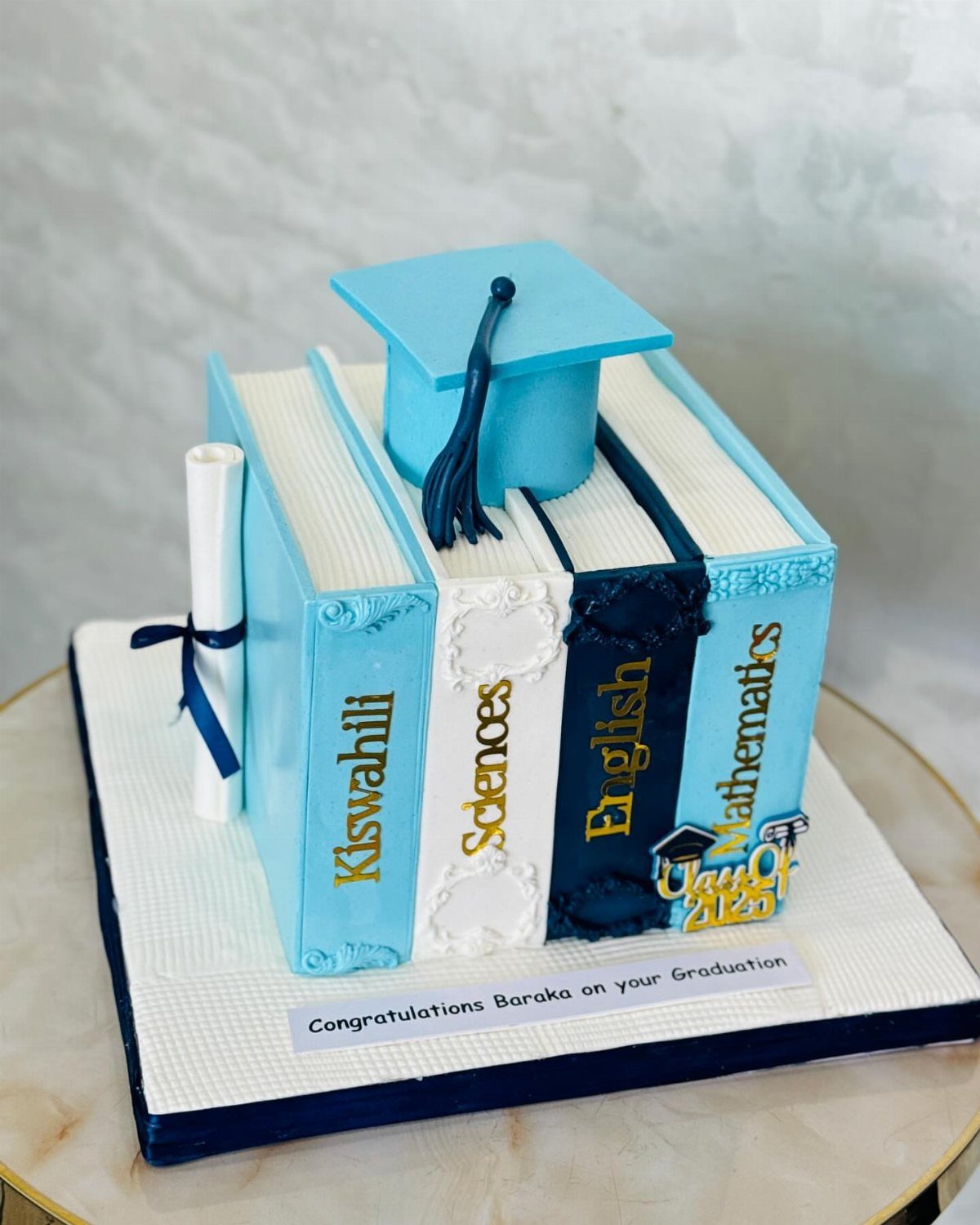 Graduation cake shaped like books with a cap, diploma, and personalized name message.