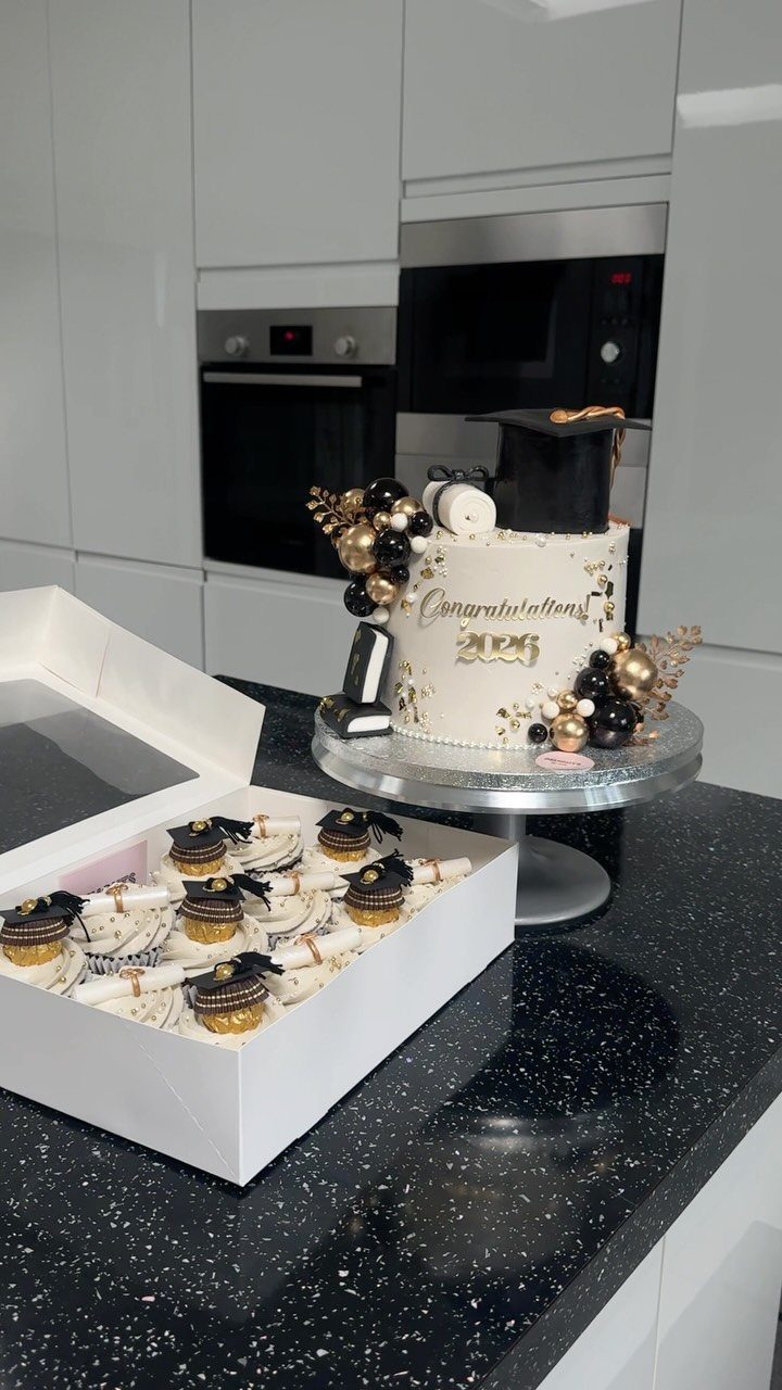 Graduation cake with a black cap and gold balloons next to graduation cupcakes.