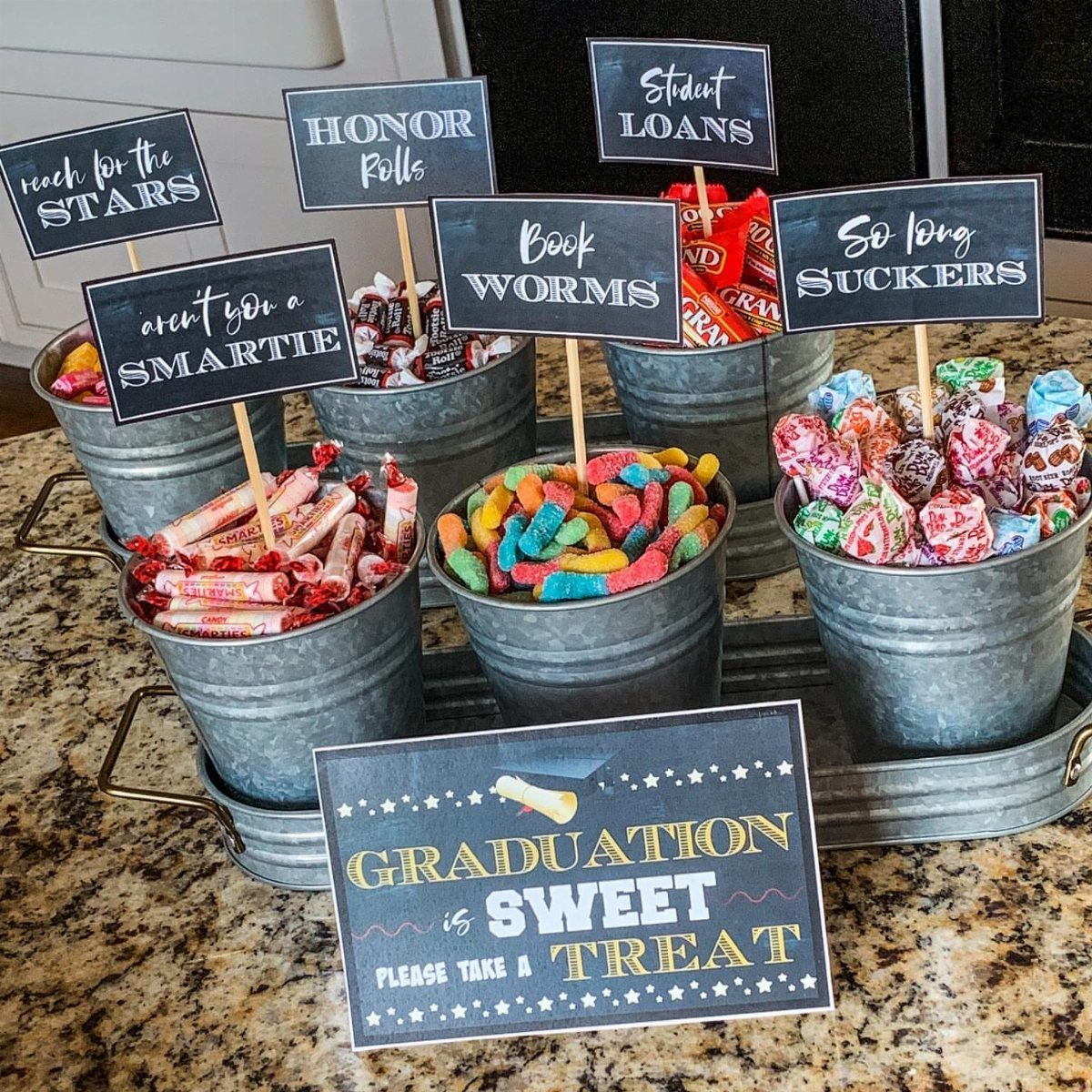 Candy bar with colorful sweets in metal buckets and graduation-themed pun signs.