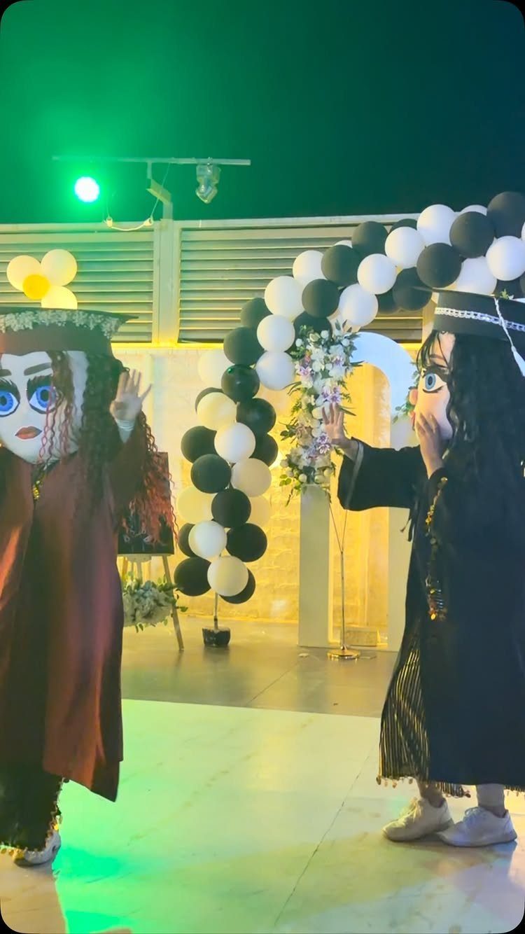 Two people in graduation mascot costumes posing by a black and white balloon arch.