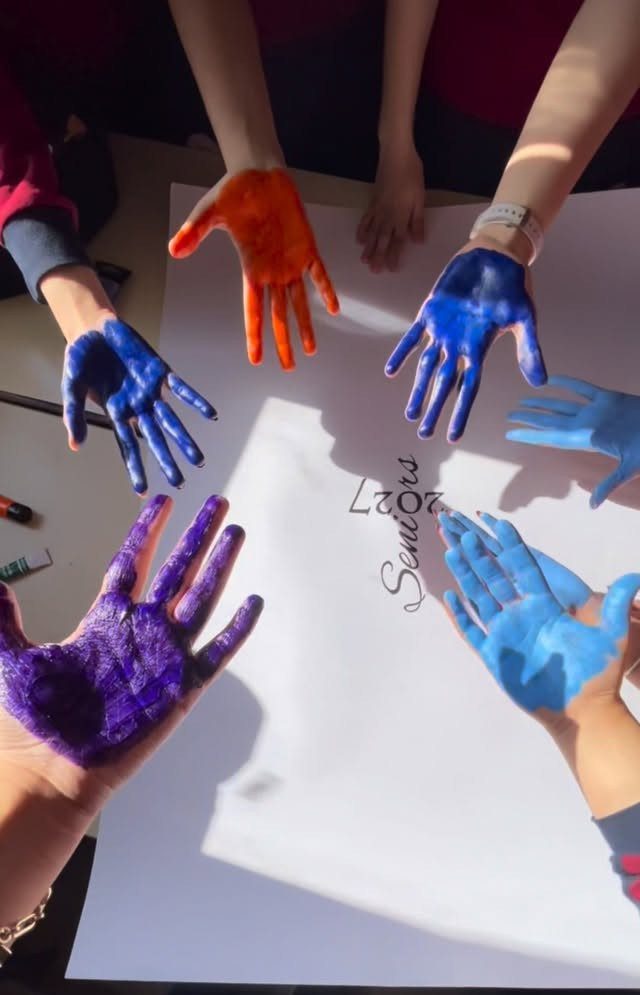 Paint-covered hands of seniors gathered around a white paper for a graduation activity.