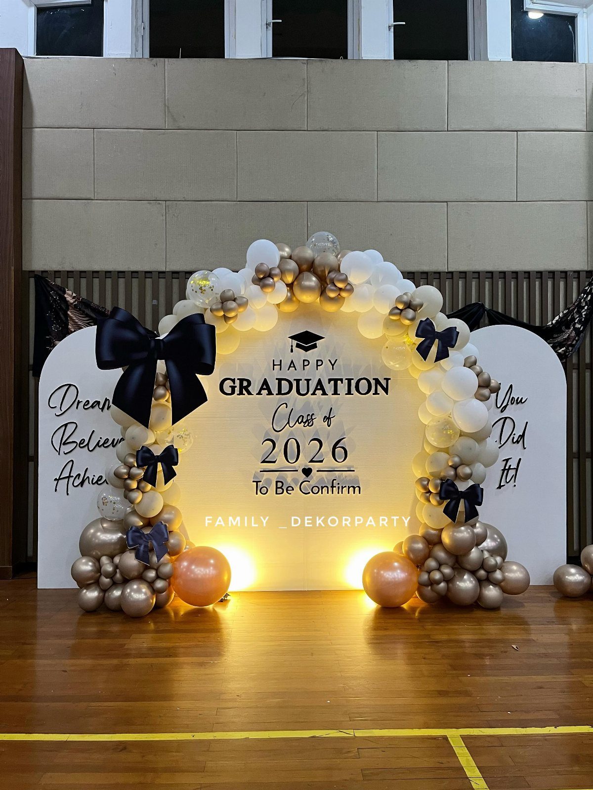 Graduation backdrop with gold and white balloons and black bows on wooden floor