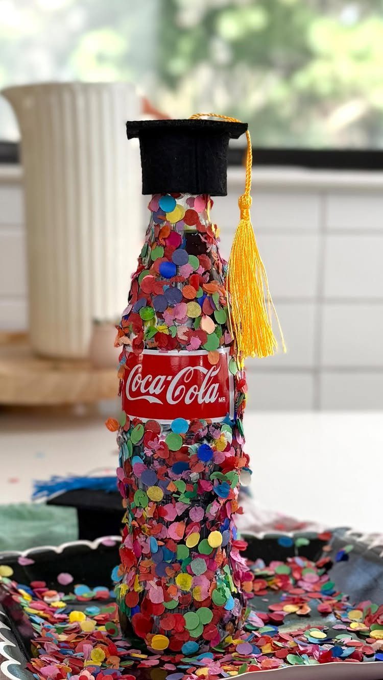 Confetti covered Coca-Cola bottle wearing a small black graduation cap with yellow tassel