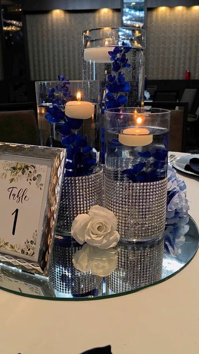 Glass vase centerpieces with blue flowers and floating candles on a mirrored base.
