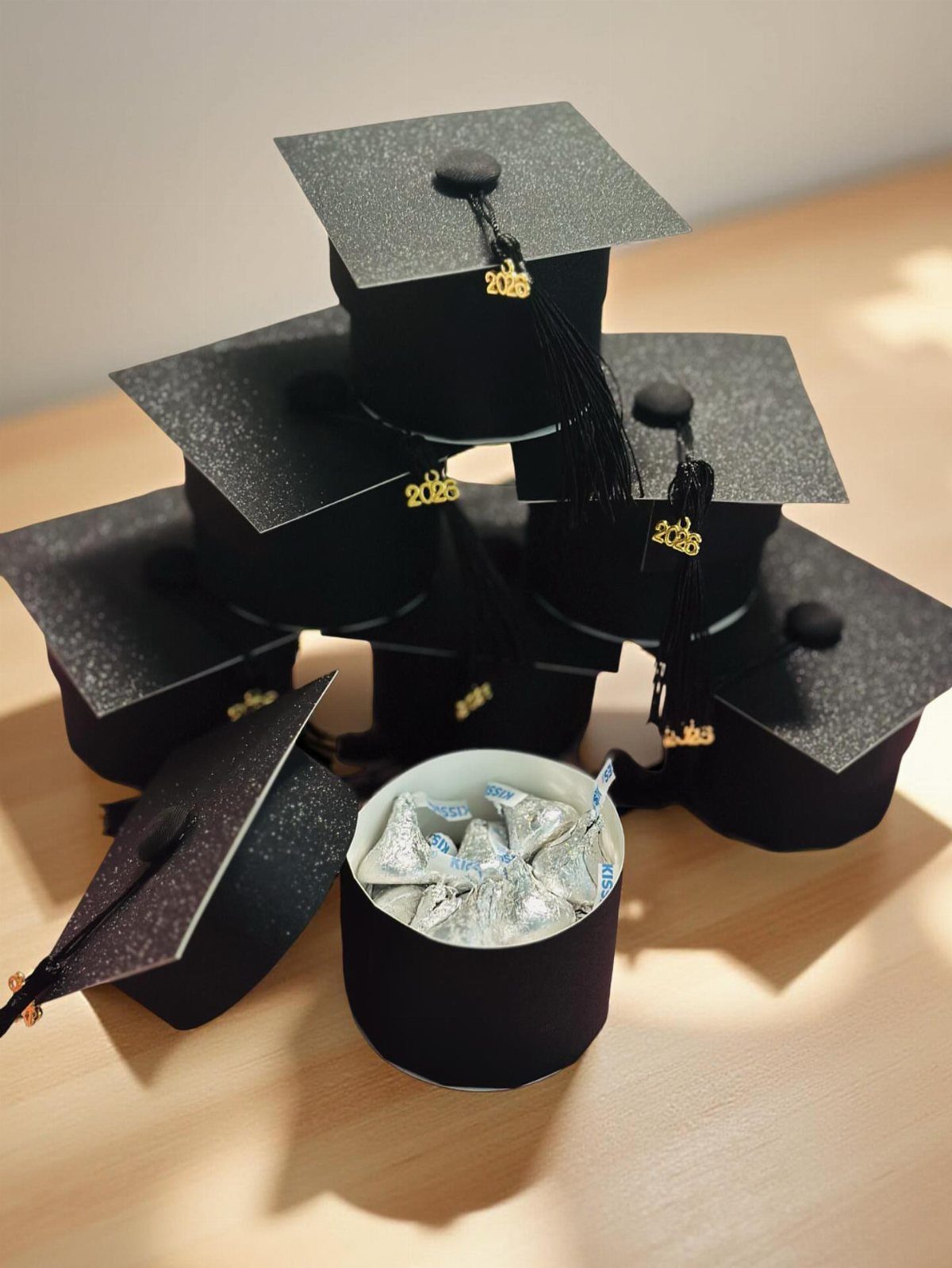 Small black glitter graduation cap containers filled with silver wrapped candies on wood