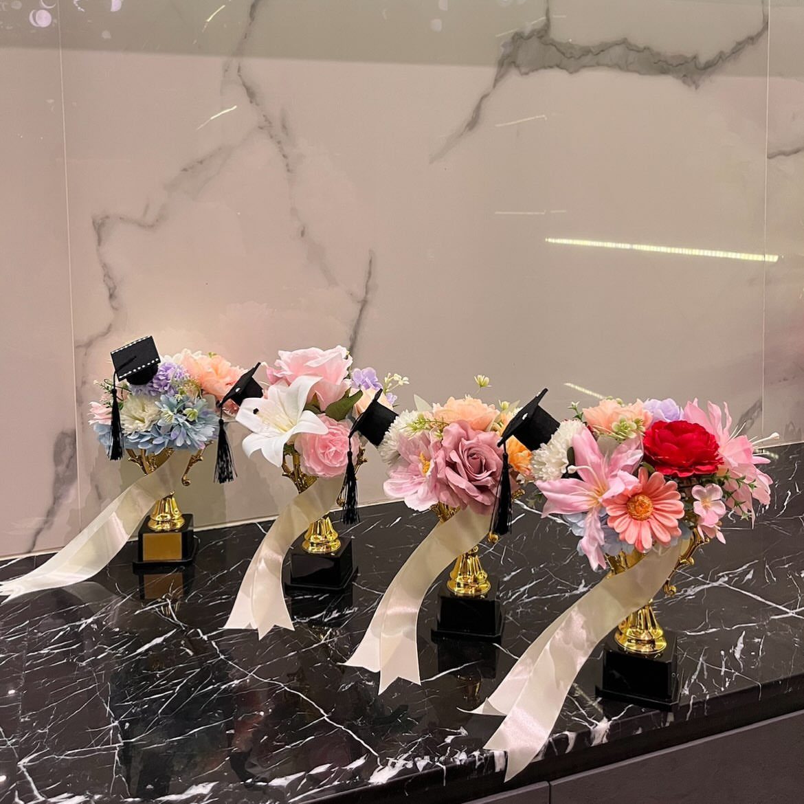 Gold trophy cups filled with colorful flowers and small graduation caps on marble.