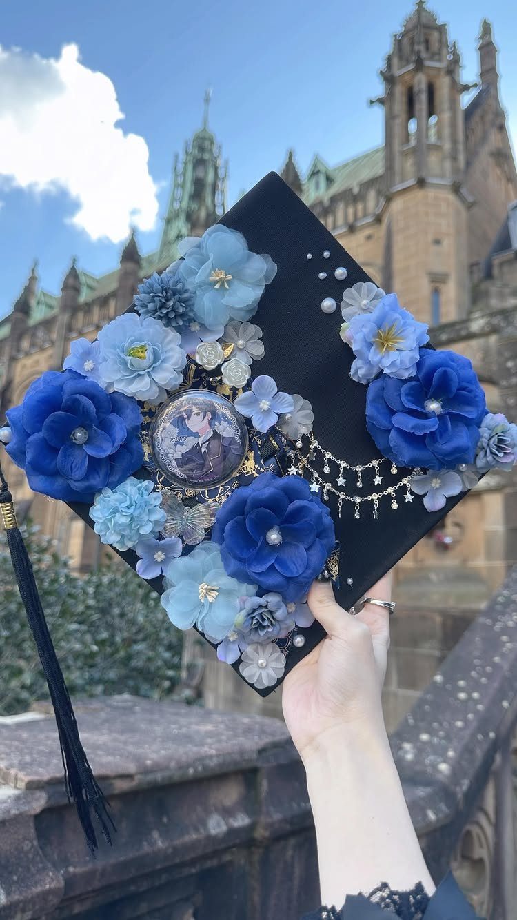 Black graduation cap decorated with blue fabric flowers, pearls, and a small portrait.