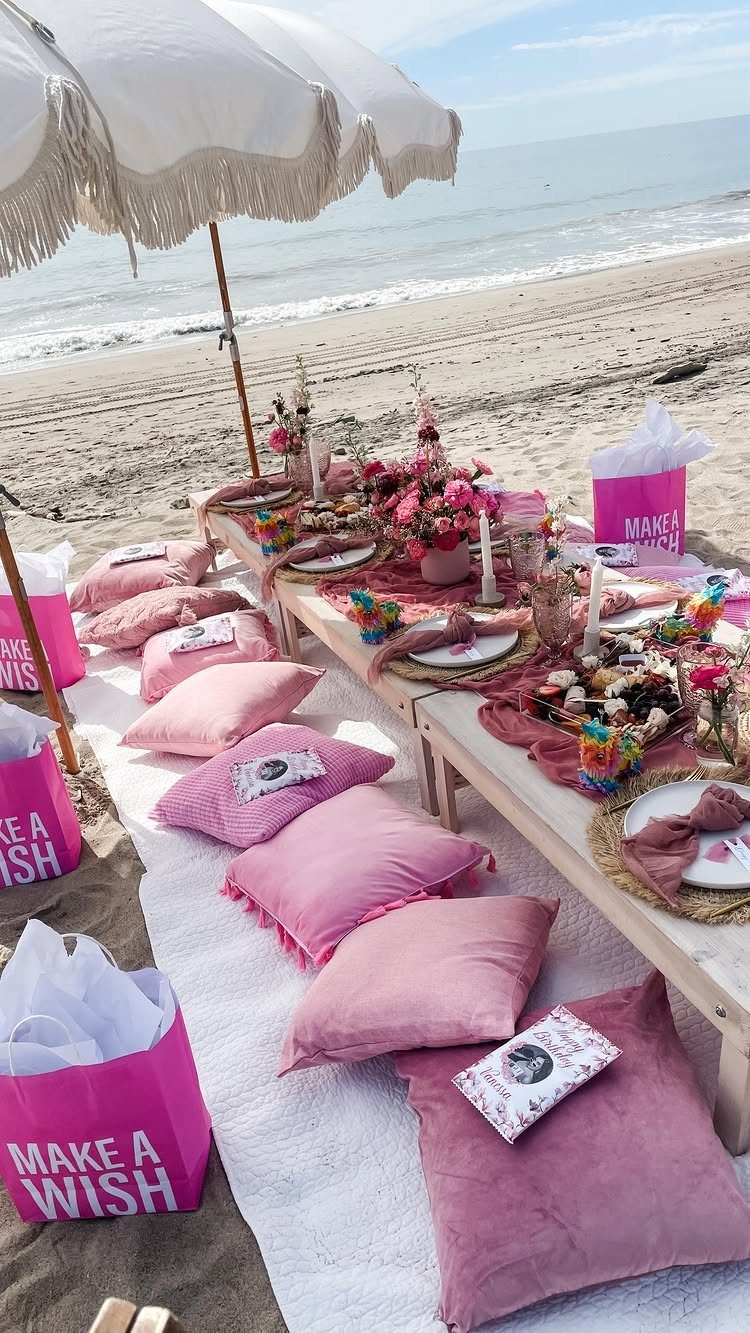 Pink beach picnic setup with low table, pillows, and 'Make a Wish' bags.