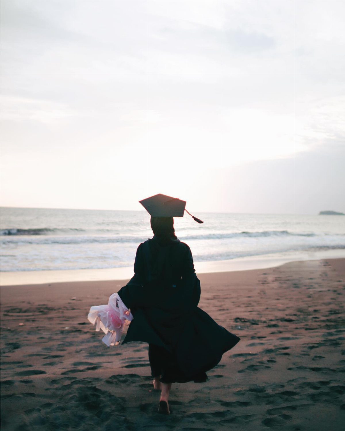 Graduate in cap and gown walking on a sandy beach toward the ocean.