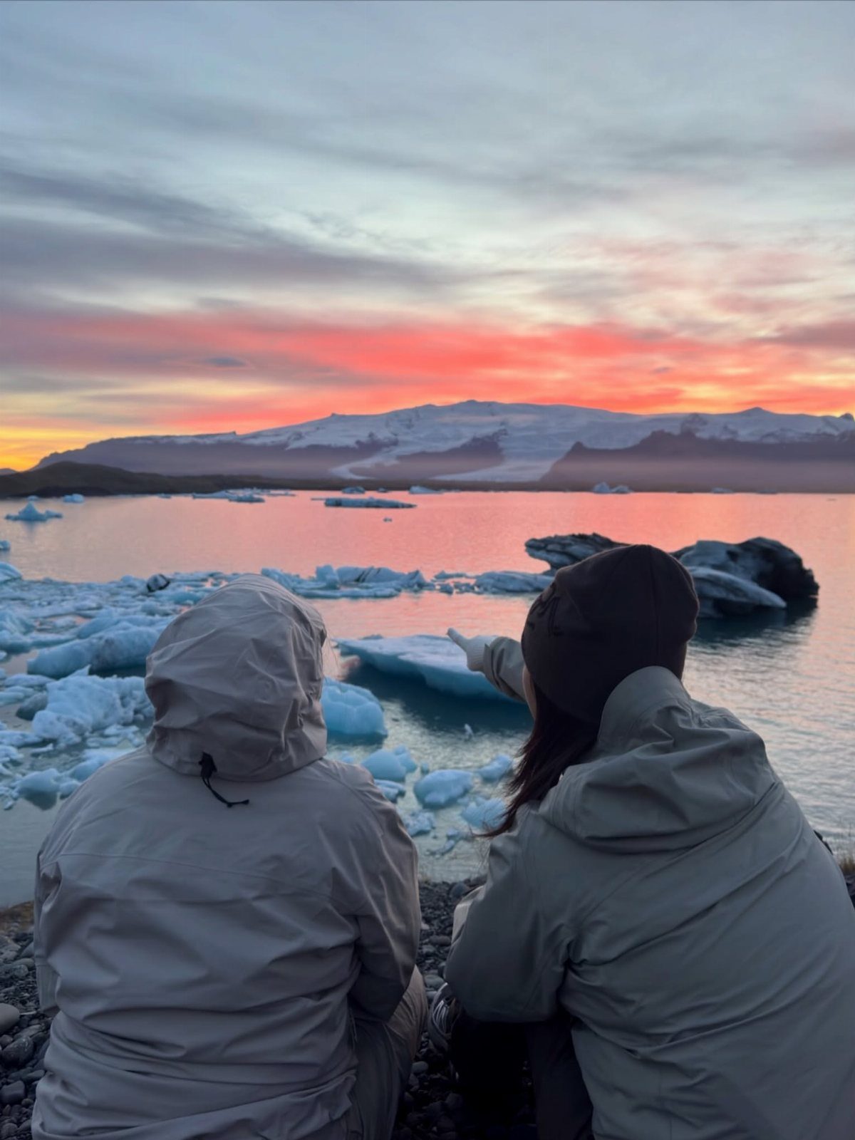 Two people in jackets watching a sunset over a glacial lagoon with icebergs.