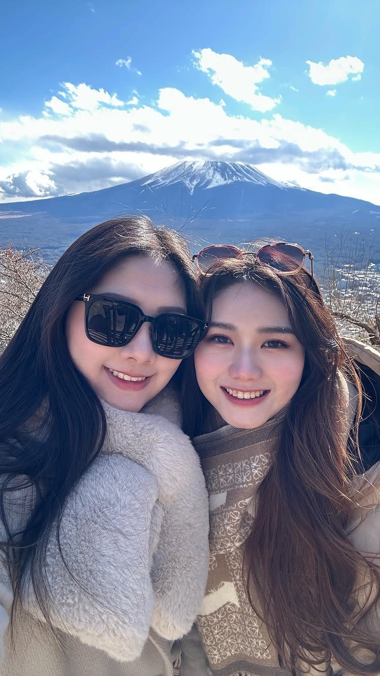 Two smiling women in winter clothes posing before a snow-capped mountain peak.
