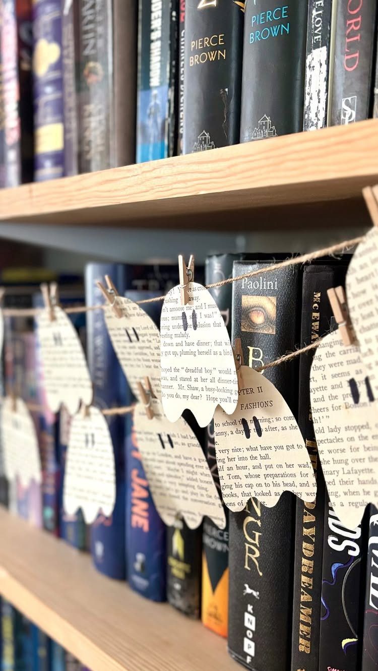Ghost-shaped book page cutouts hanging from a twine garland on a bookshelf.