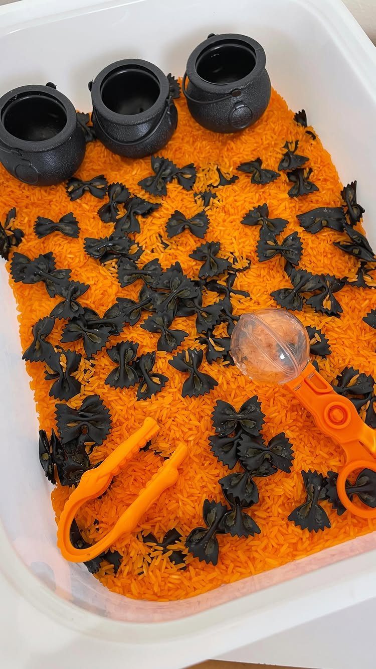 Sensory bin with orange rice, black bow-tie pasta, and small black cauldrons.