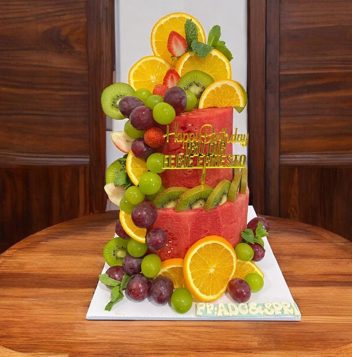 Watermelon cake decorated with fresh orange slices, kiwi, grapes, and a gold topper.