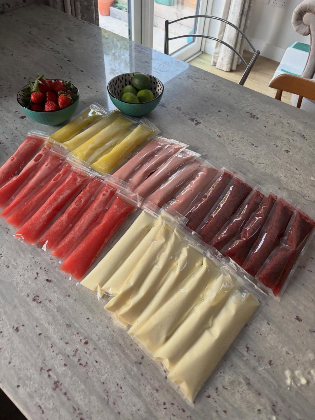 Colorful frozen ice pops in plastic sleeves on a marble countertop with fresh fruit.