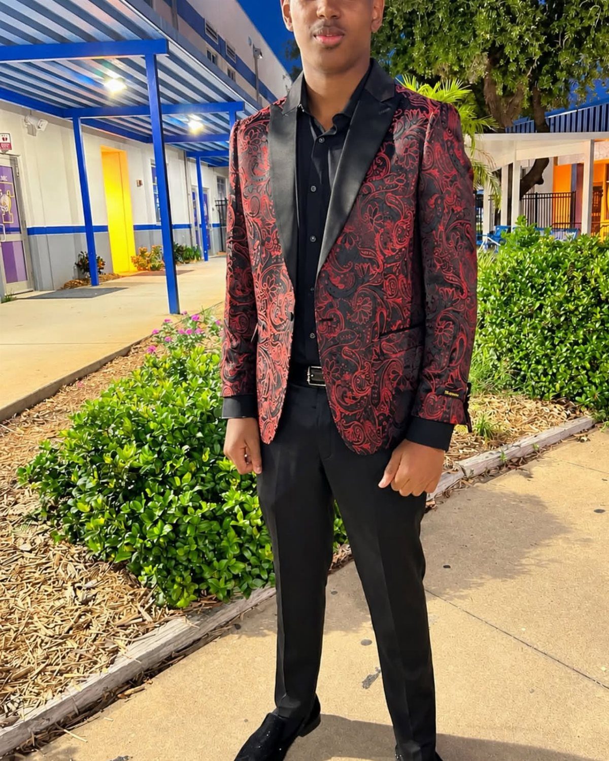 Young man wearing a red and black paisley blazer and black trousers outdoors.
