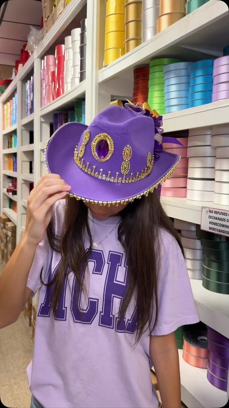 Person wearing a decorated purple cowboy hat and matching purple t-shirt in a store.