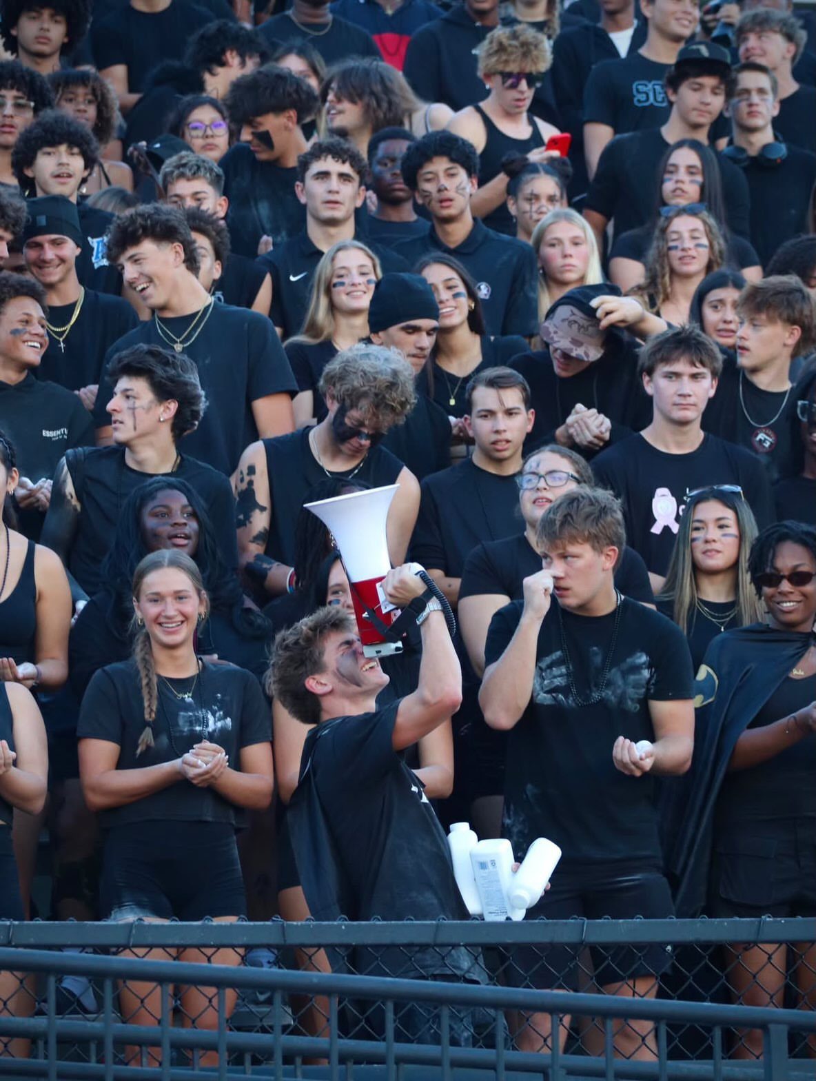 Teenagers wearing all black clothing and face paint cheering in stadium bleachers.