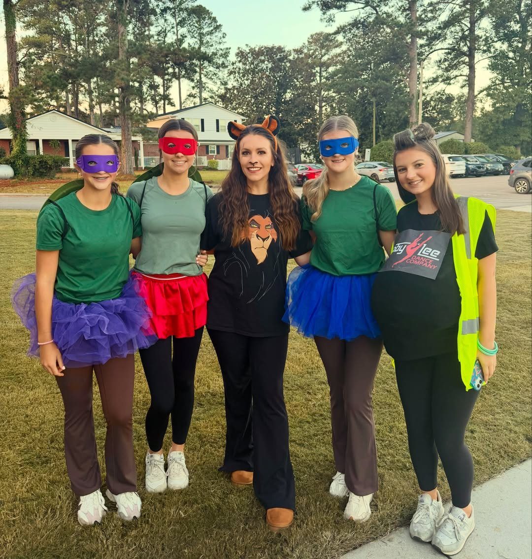 Five teenage girls in colorful superhero costumes and masks posing outdoors on grass.