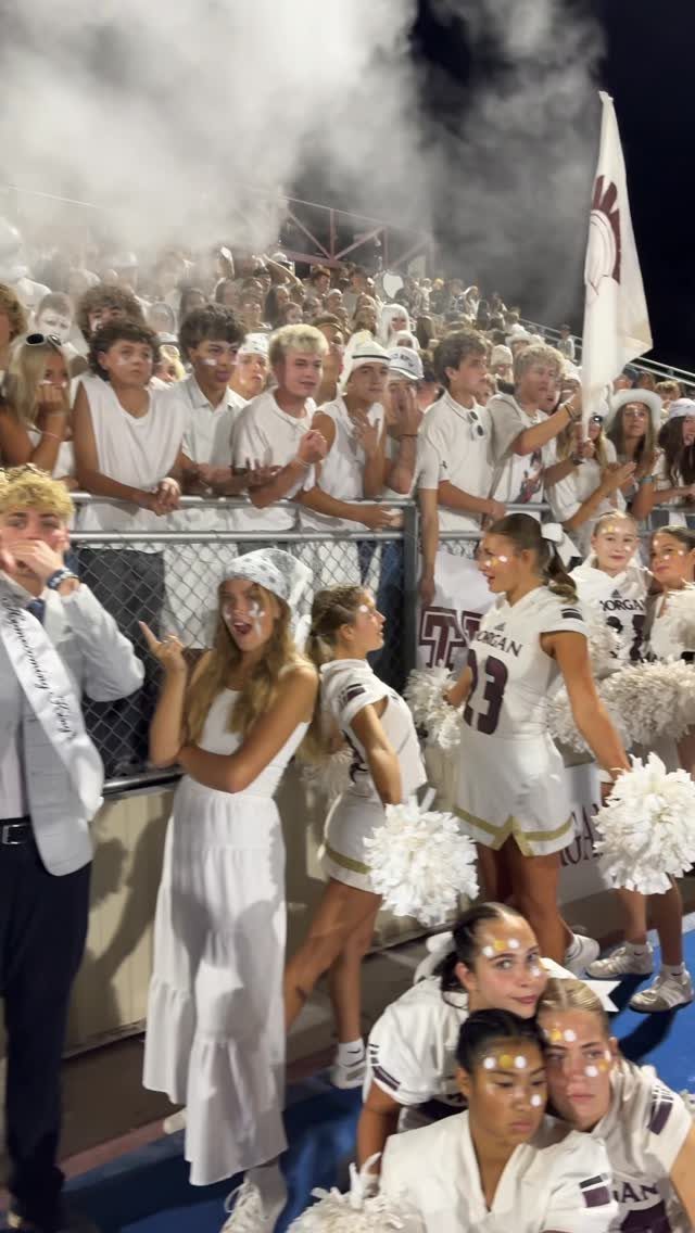 Students and cheerleaders in white outfits at a night game with smoke background.