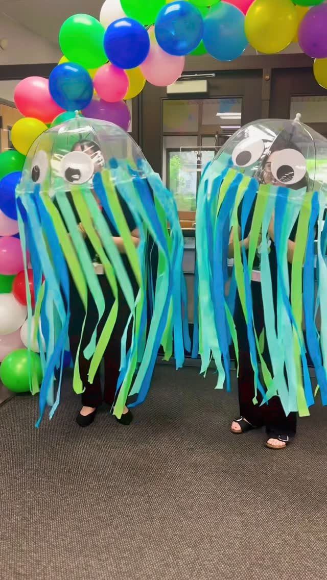 Two people in homemade jellyfish costumes standing before a colorful balloon arch.