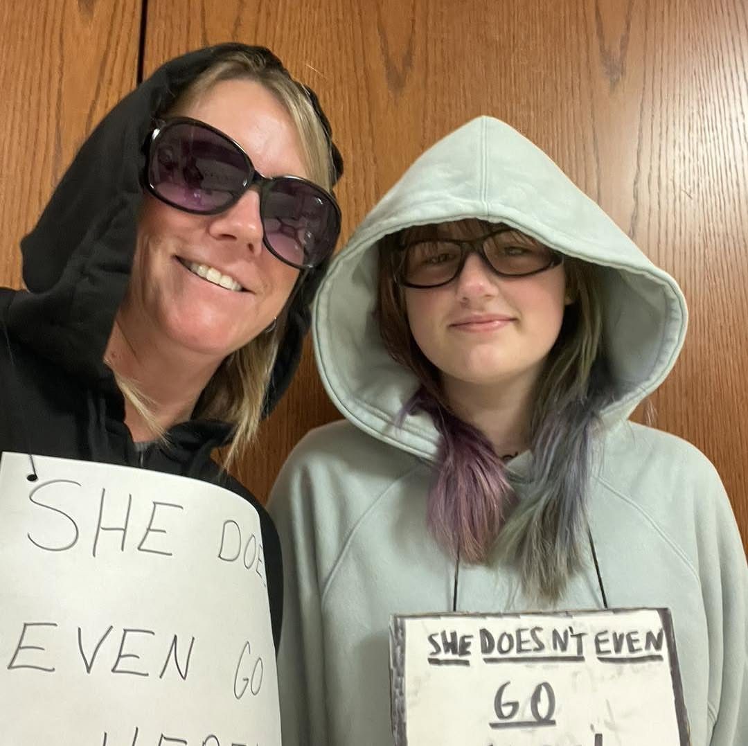 Two women wearing hoodies and sunglasses holding signs saying she doesn't even go here