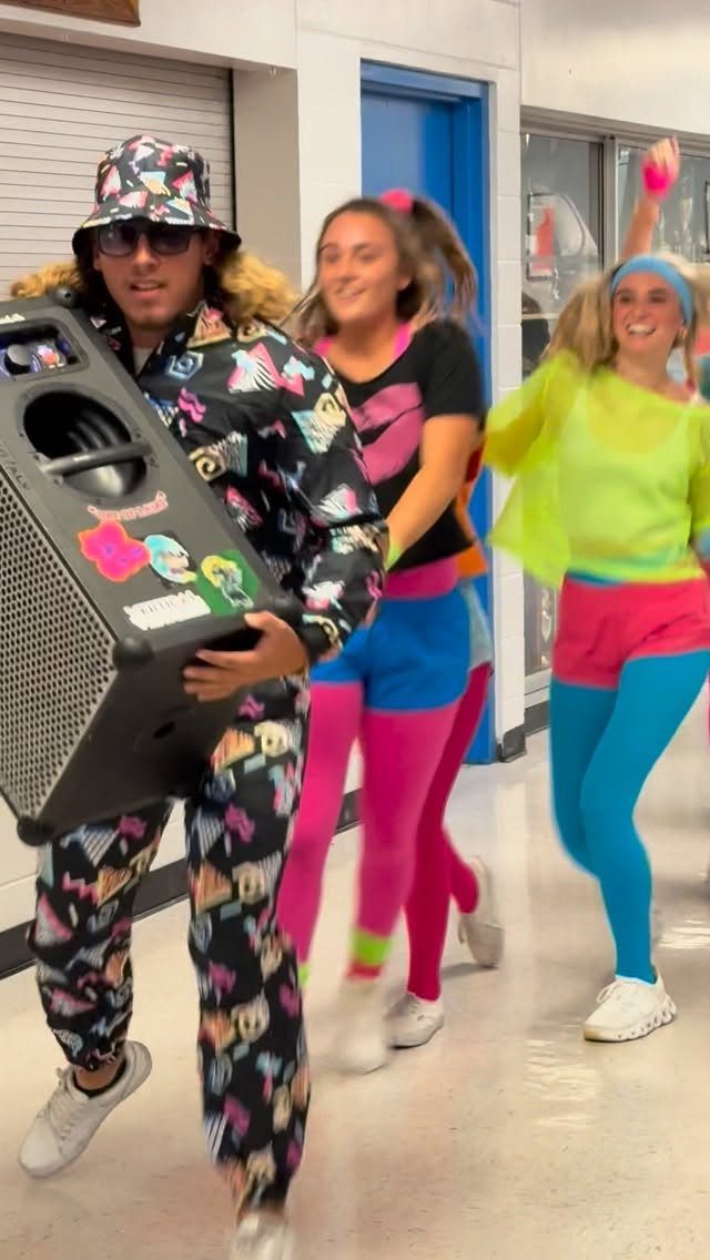 Teenagers in neon 80s outfits walking through a school hallway with a speaker