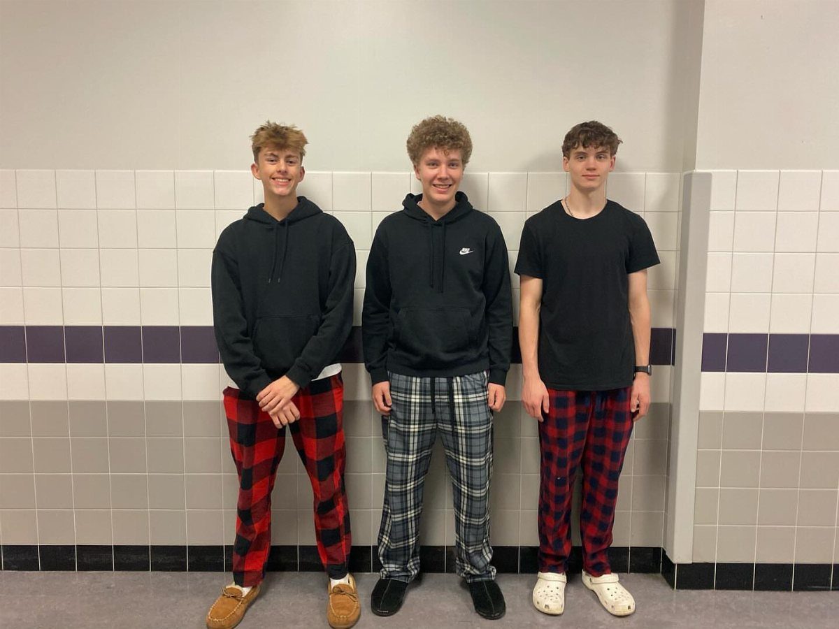 Three teenage boys wearing black tops and plaid pajama pants in a school hallway.