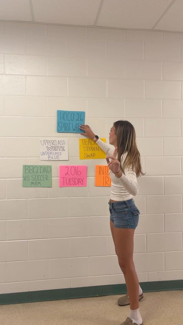 Young woman pinning colorful spirit week theme signs to a white brick wall