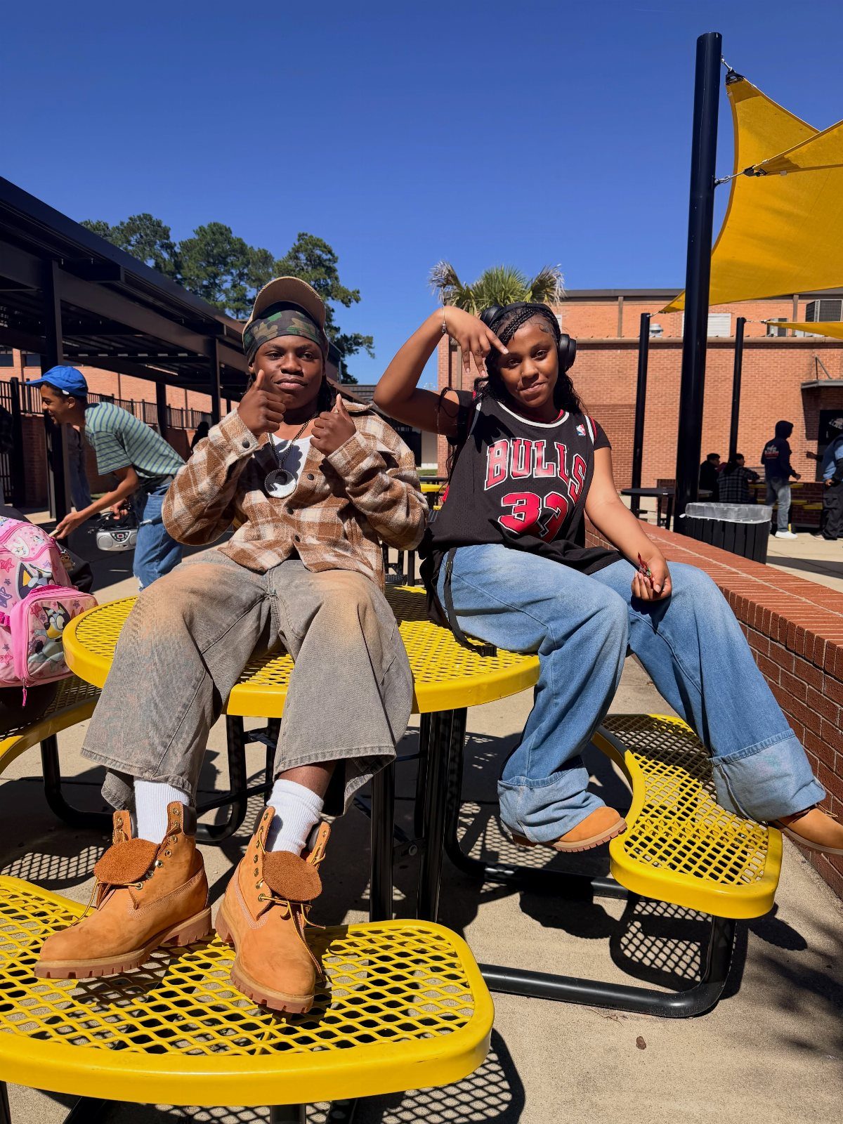Two teenagers wearing retro 90s style clothing posing on yellow outdoor school tables.