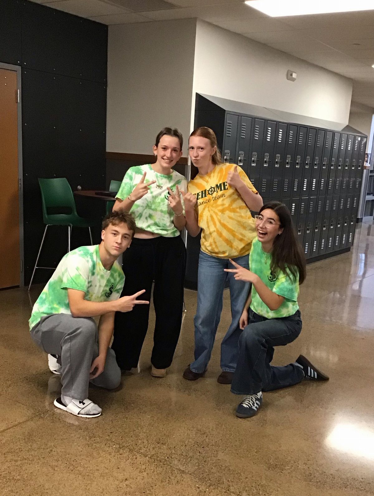 Four students wearing colorful tie-dye shirts posing in a school hallway by lockers.