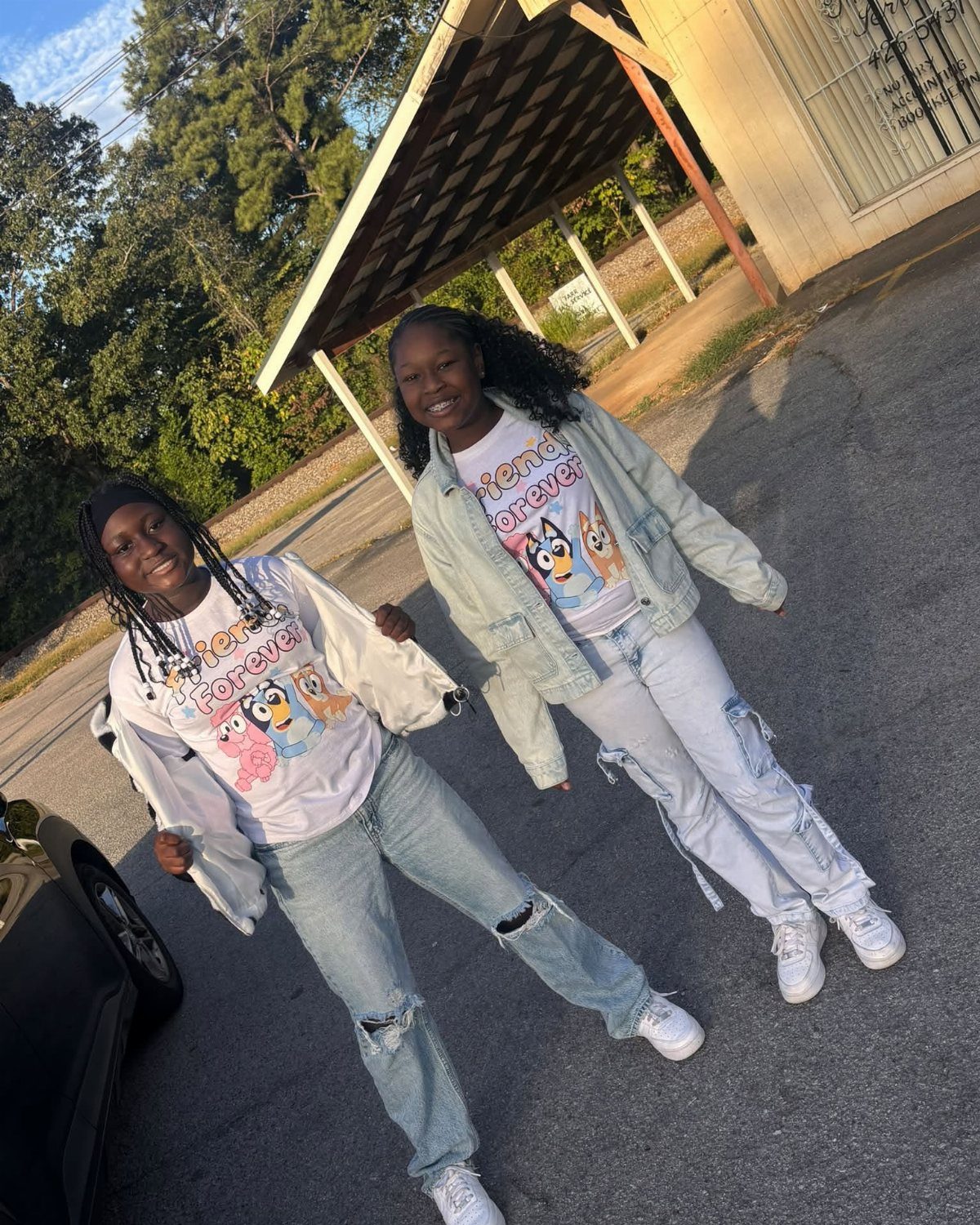 Two young women wearing matching graphic t-shirts and ripped jeans posing outdoors together.