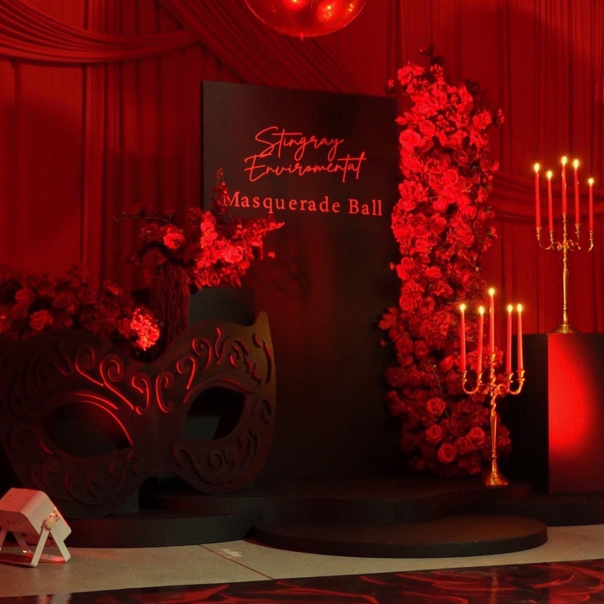 Masquerade ball decor with gold candelabras, a large black mask, and red roses.