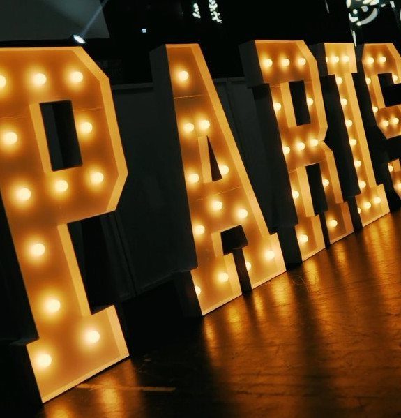 Large glowing marquee letters spelling PARIS on a polished wooden floor