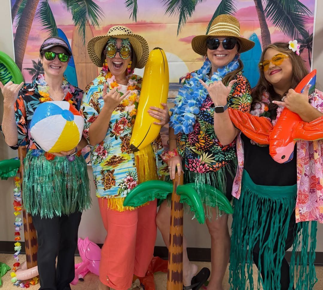 Four women in tropical shirts and leis posing with beach inflatables and backdrop.