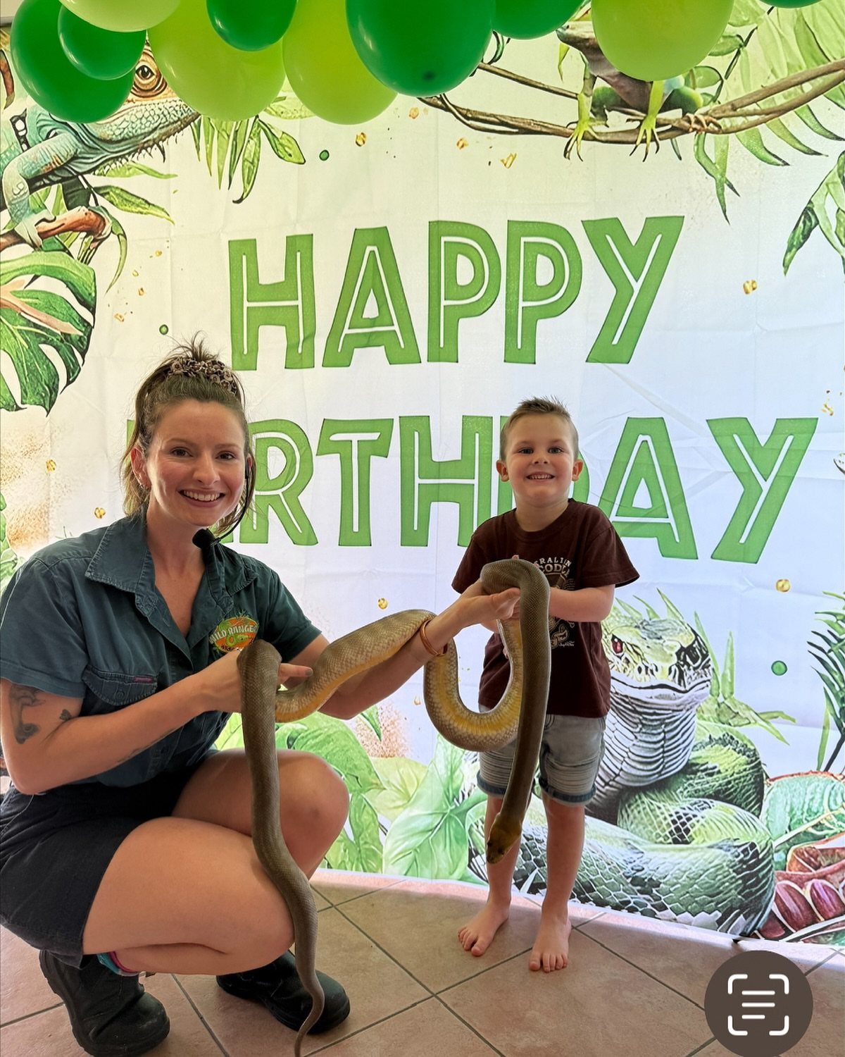 Woman and young boy holding a large brown snake in front of birthday backdrop