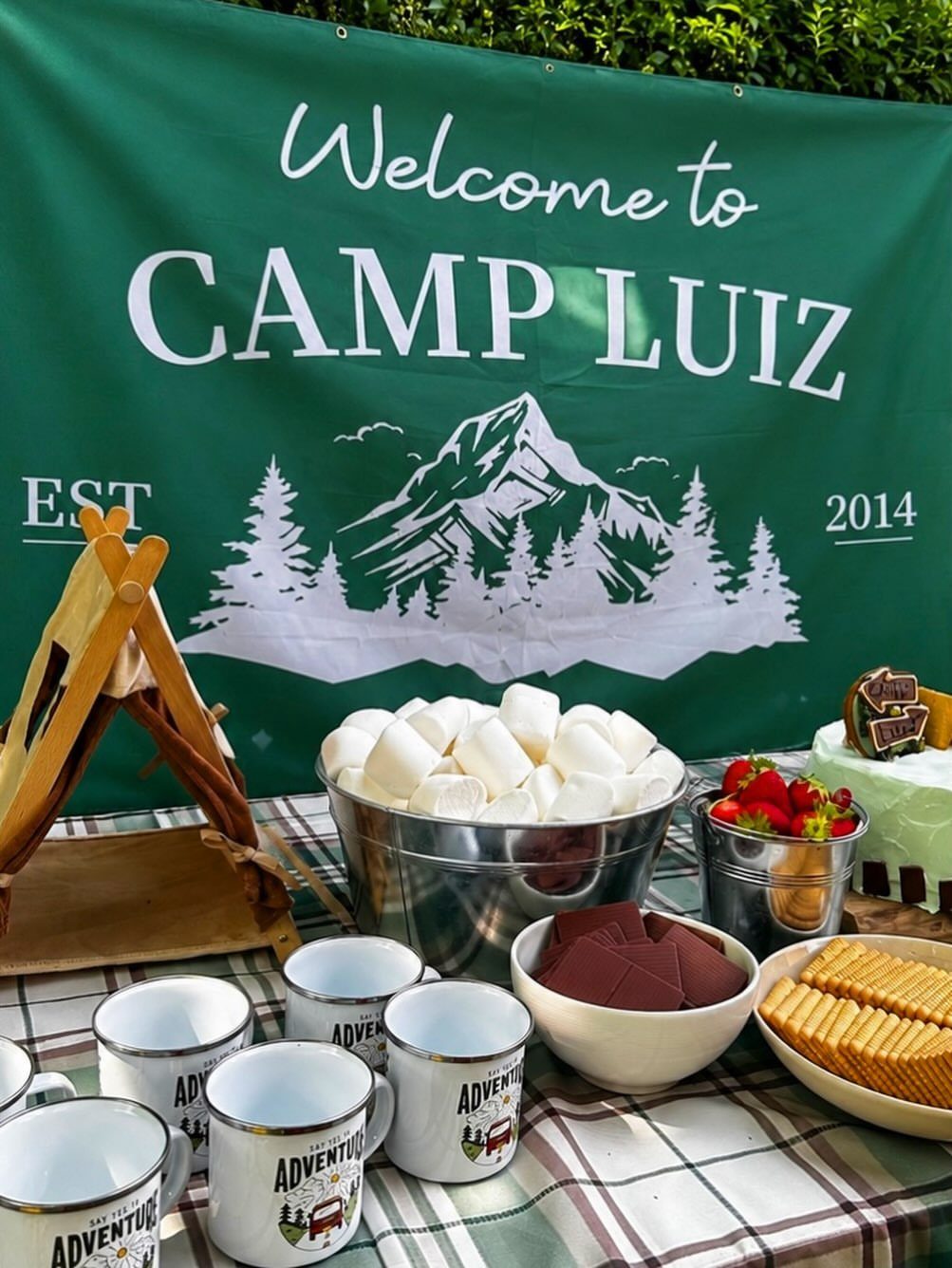 Camping themed party table with a Camp Luiz banner, marshmallows, and mini teepee.