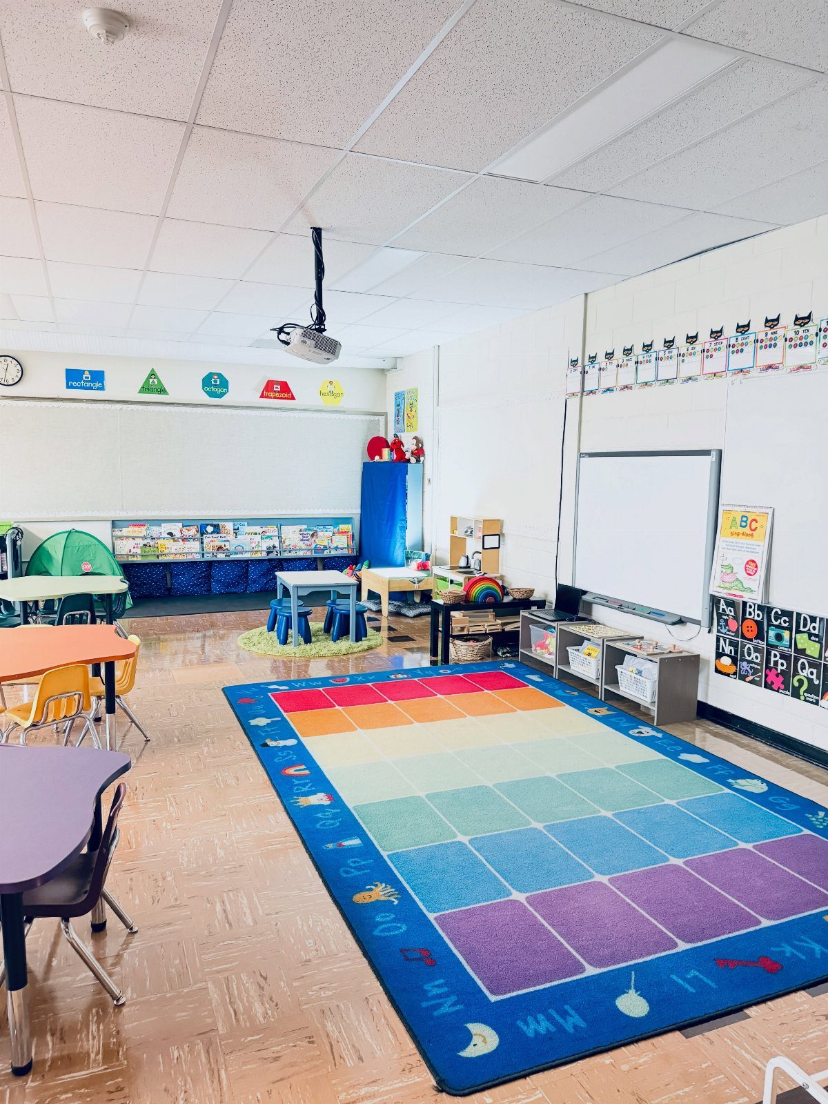 Colorful kindergarten classroom featuring a rainbow rug, small tables, and educational wall displays.