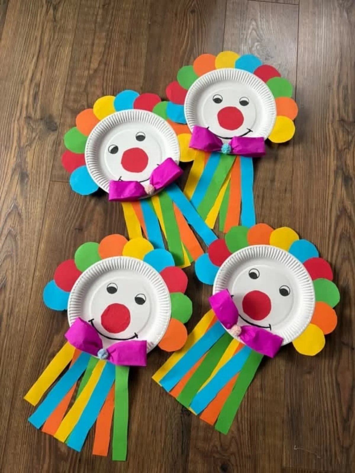 Four colorful clown faces made from paper plates and construction paper on wood.