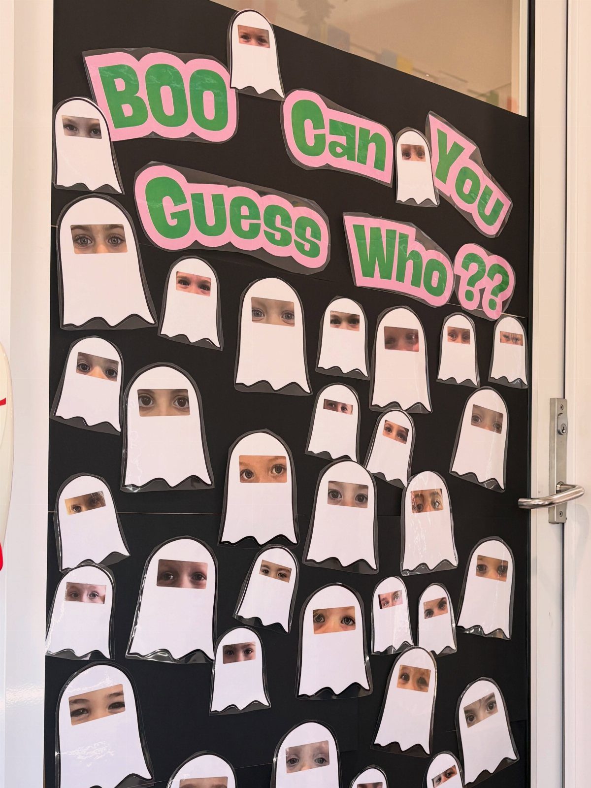 Black bulletin board with white paper ghosts and children's eyes for a guessing game.