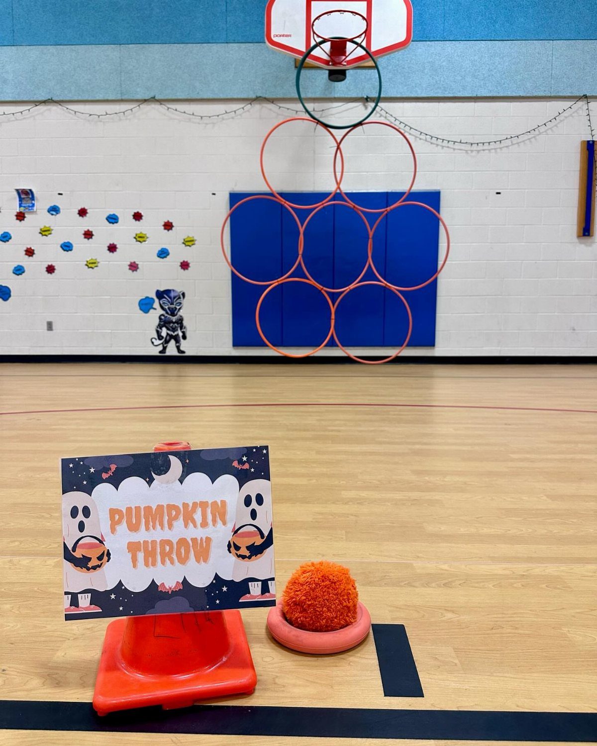 Pumpkin Throw game setup with a sign, plush pumpkin, and hula hoops in gym.