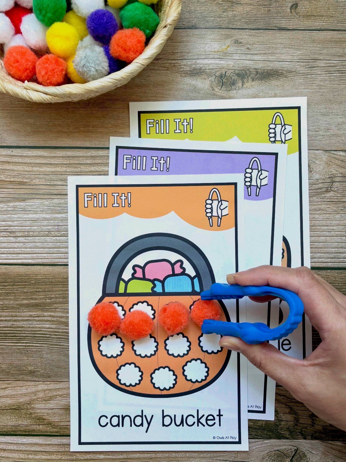 Hand using blue tweezers to place orange pom-poms on a candy bucket mat.