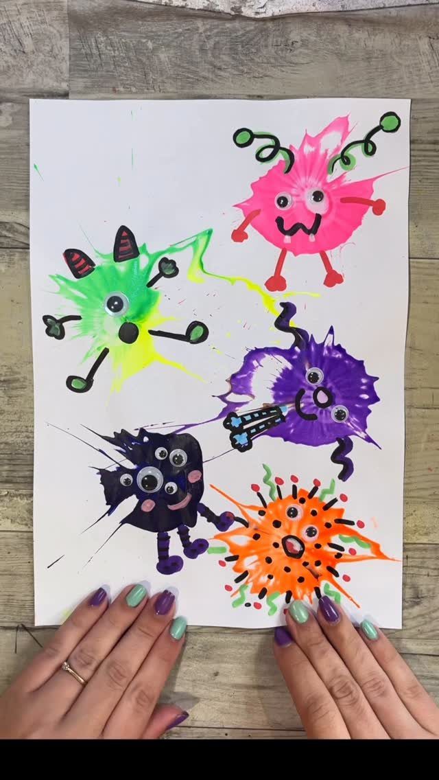 Colorful paint splatter monsters with googly eyes on white paper held by hands.
