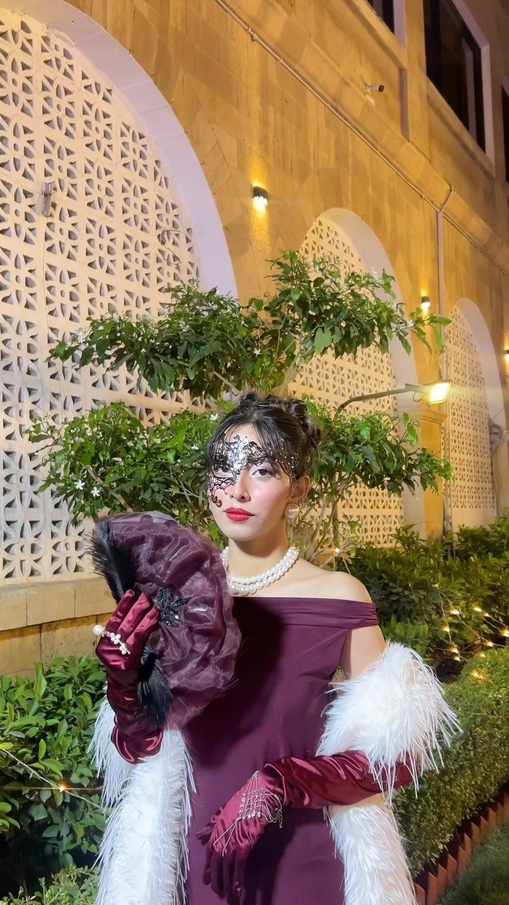 Woman in burgundy gown and black lace mask holding a feathered fan.
