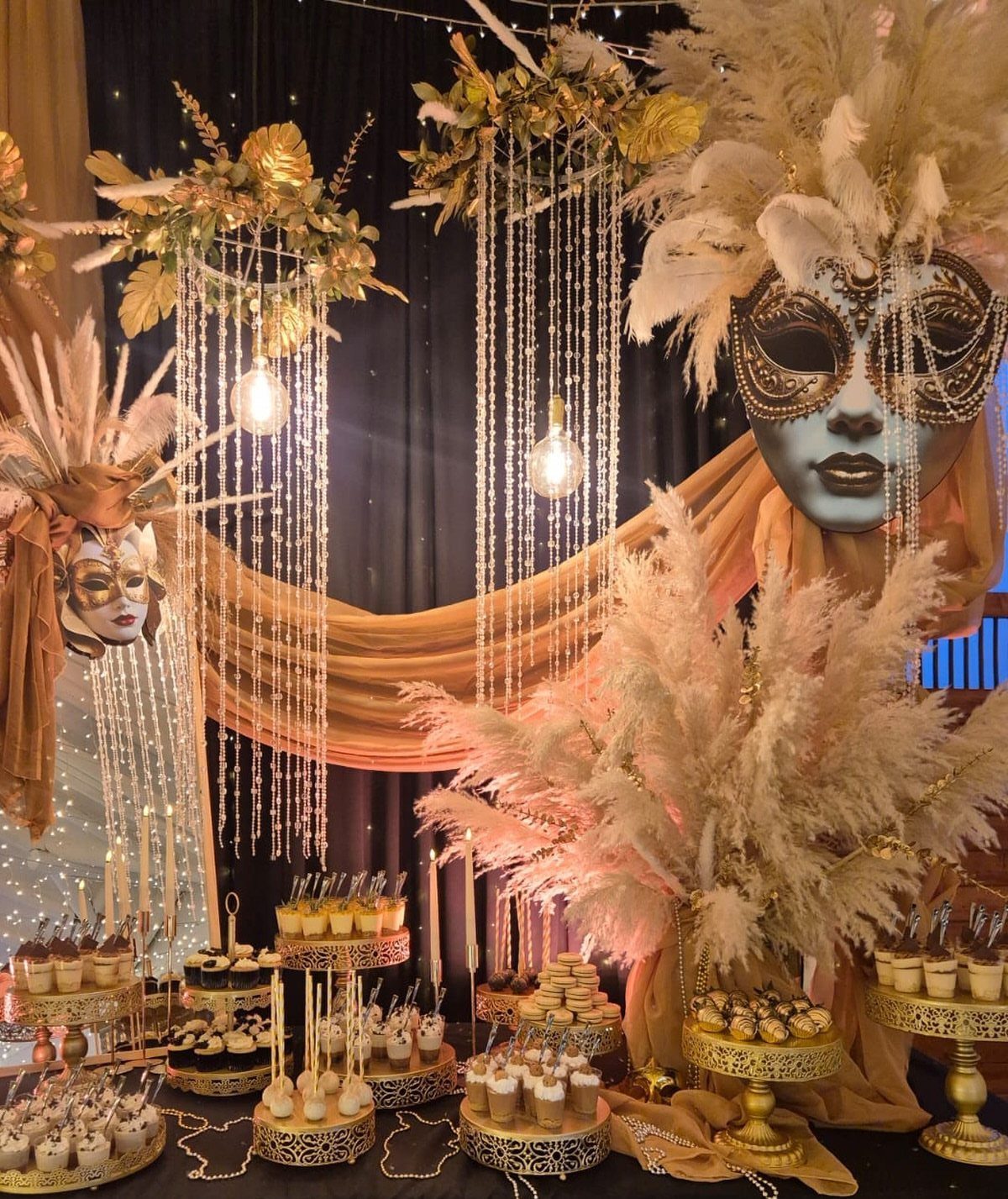 Gold fabric draping and large masquerade masks decorating a lavish dessert table setup.