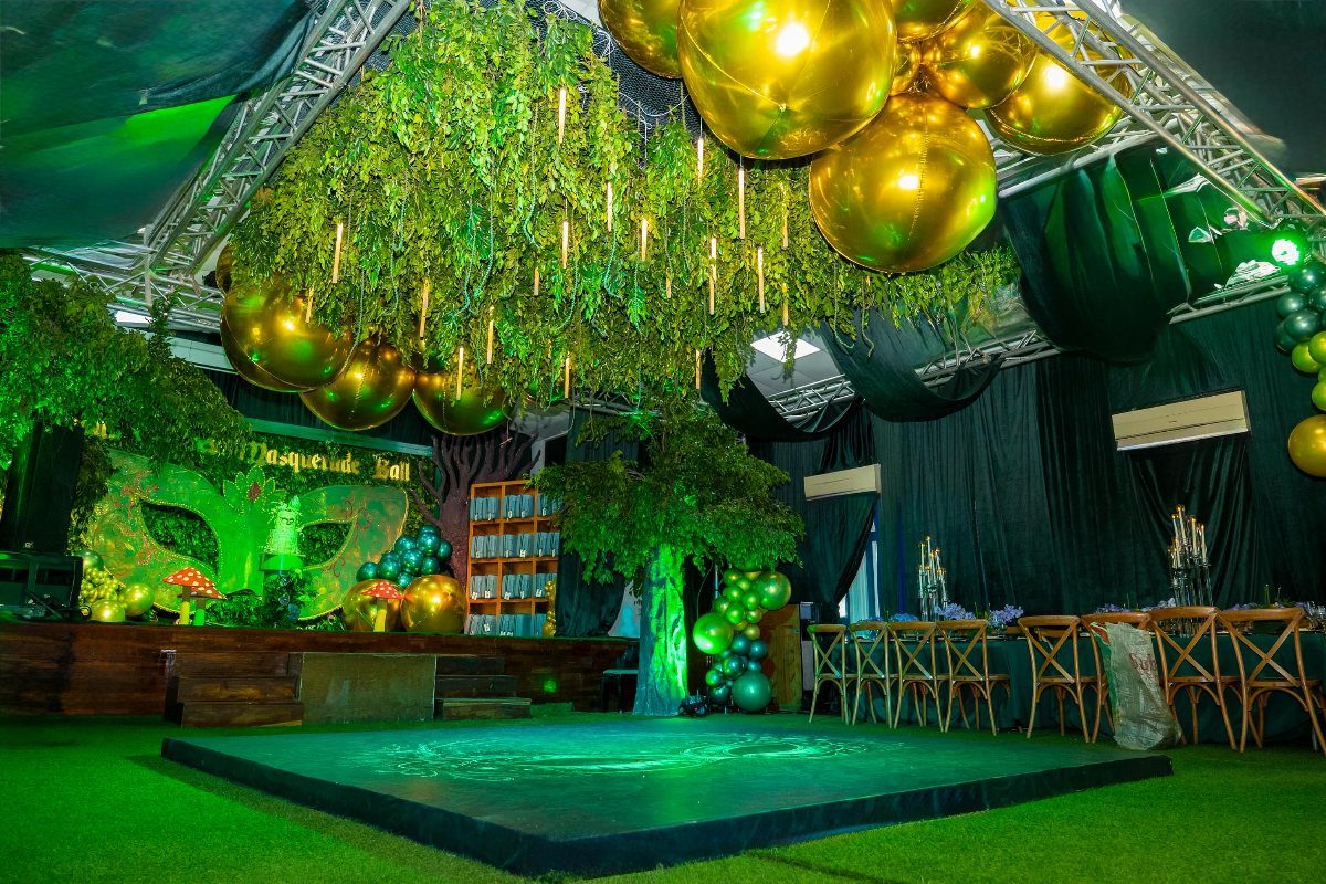 Event space with hanging green foliage, gold balloons, and green lighting for masquerade.
