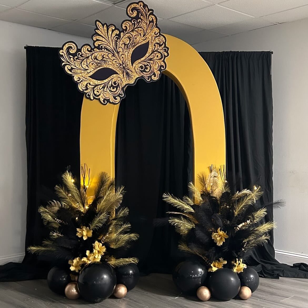 Gold arched backdrop with a large ornate mask and black feather arrangements.